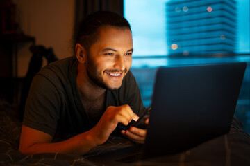 Portrait of young smiling man holding smartphone in hand, working at laptop while lying on the bed close to evening time.