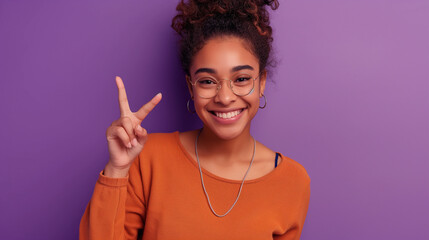 Portrait of a cheerful young woman gesturing peace sign over on purple color background...