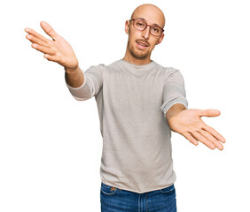 Bald man with beard wearing casual clothes and glasses looking at the camera smiling with open arms...