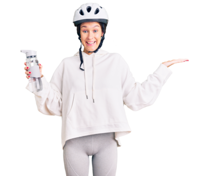 Beautiful brunette young woman wearing bike helmet and holding water bottle celebrating victory with happy smile and winner expression with raised hands