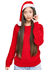 Young chinese woman wearing christmas hat mouth and lips shut as zip with fingers. secret and...