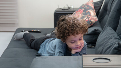 Criança com cabelos cacheados com expressão pensativa brincando de jogos no celular deitado no sofa, pensativo, ideia - obrazy, fototapety, plakaty