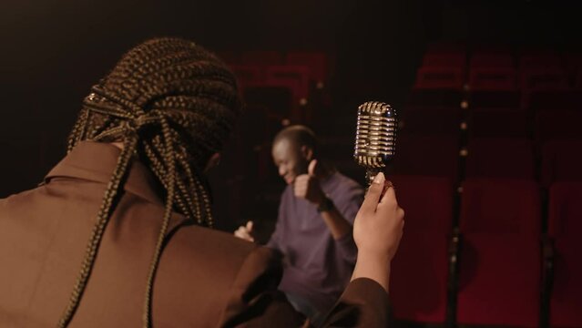 Back view shot of young Black woman standing with retro mic on stage at amateur theater and giving standup concert to her colleague sitting in first row and clapping