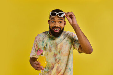A man wearing a colorful tie-dye shirt is holding a drink in his hand, standing in a relaxed pose.