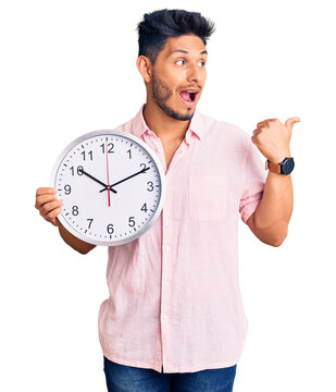 Handsome latin american young man holding big clock pointing thumb up to the side smiling happy with open mouth