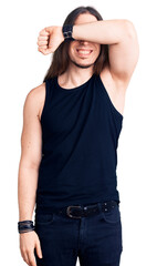 Young adult man with long hair wearing goth style with black clothes covering eyes with arm smiling cheerful and funny. blind concept.