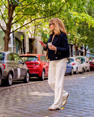 Full length of an attractive mid aged woman holding paper coffee cup and her mobile phone in her hand and walking on the city street