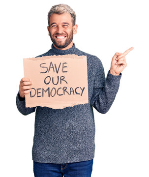 Young blond man holding save our democracy cardboard banner smiling happy pointing with hand and finger to the side