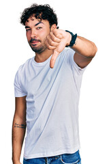 Hispanic young man with beard wearing casual white t shirt looking unhappy and angry showing rejection and negative with thumbs down gesture. bad expression.