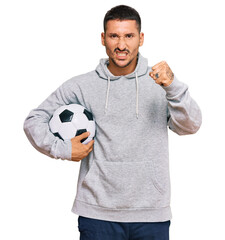 Handsome man with tattoos holding soccer ball annoyed and frustrated shouting with anger, yelling crazy with anger and hand raised