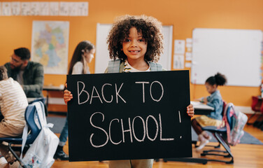 Proud pupil standing in a co-ed classroom on first day of elementary school, holding a back to school sign - 774980244