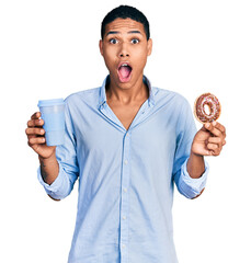 Young hispanic man eating doughnut and drinking coffee afraid and shocked with surprise and amazed...