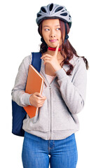 Young beautiful chinese girl wearing student backpack and bike helmet holding book serious face thinking about question with hand on chin, thoughtful about confusing idea