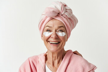 Skincare. Very happy old Caucasian woman laughing, applying cosmetic eye patches mask, reduces wrinkles, wears wrapped towel on head, isolated on white background. F