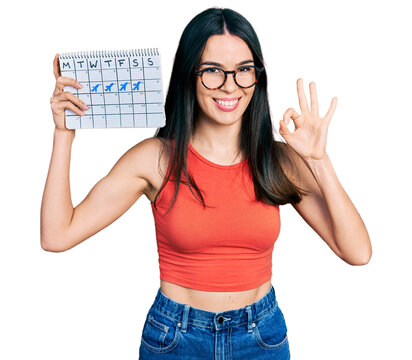 Young hispanic woman holding travel calendar doing ok sign with fingers, smiling friendly gesturing excellent symbol