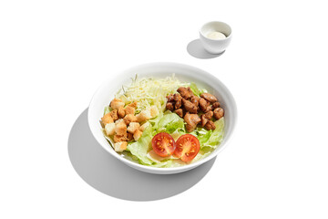 Fresh Caesar salad with crispy croutons and grilled chicken pieces in a white bowl, isolated on a white background with dressing on the side