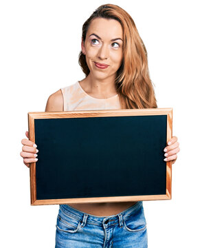 Young caucasian woman holding blackboard smiling looking to the side and staring away thinking.