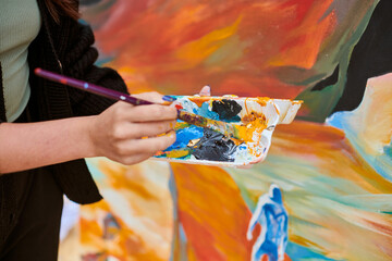 Female painter hand dips paintbrush into palette of colorful paints for live painting of picture for outdoor street exhibition, close up view of female artist hand holding paintbrush - Powered by Adobe