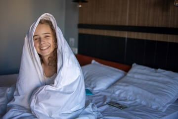 A charming portrait of a blonde teenage girl in a white bedspread, sitting on a large comfortable...