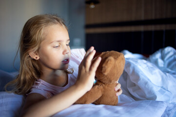 A little blonde girl in cute pink pajamas is playing with a plush brown teddy bear on a large bed...