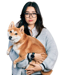 Beautiful hispanic woman holding cute dog relaxed with serious expression on face. simple and natural looking at the camera. - obrazy, fototapety, plakaty