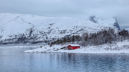 Small red wooden house on the white snowy lakeshore seen from the lake in Norway