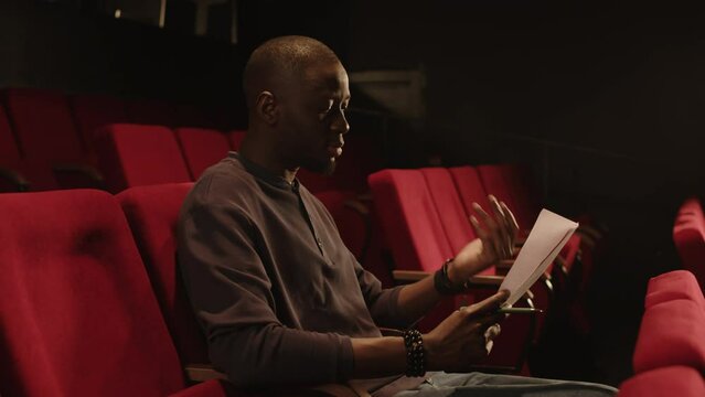 Medium shot of young expressive Black male actor sitting on red seat and revising monologue during auditions in theater