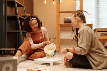 Young woman with bucket pouring white paint in tray while sitting on the floor and looking at girl paintroller during cafe renovation work - 775034819
