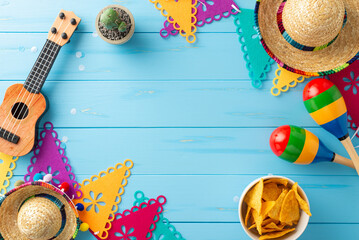 Mexican-themed arrangement for Cinco de Mayo. Overhead shot displaying cultural items: sombreros, vihuela, maracas, cactus, flag garlands, and nachos on blue wooden surface. Ideal for event promotions