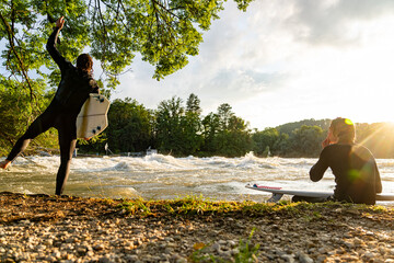 Two surfers at Aare River enjoy sunset, one balancing on rocks.