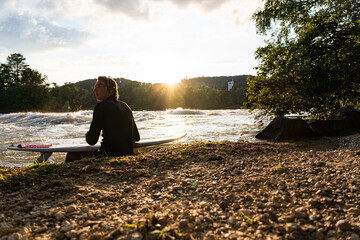 The young surfer sits by the Aare River, gazing at the sunset, enjoying the calm ambiance.