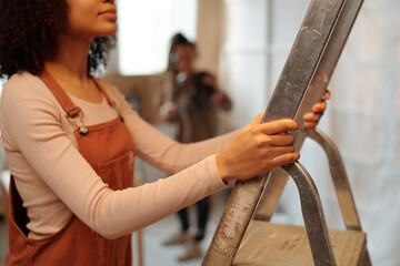 Cropped shot of young African American woman in long sleeve pullover and brown overalls holding by metallic stepladder during refit work - 775041229