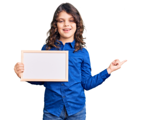 Cute hispanic child with long hair holding empty white chalkboard smiling happy pointing with hand and finger to the side