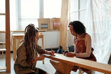 Young businesswoman with dreadlocks making notes in copybook while her colleague measuring length and width of wooden bench - 775042282
