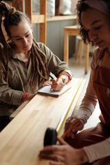 Young serious woman with dreadlocks looking at her colleague with measuring tape taking measures of wooden bench or board - 775043677