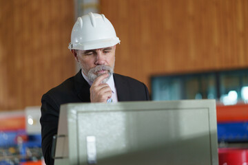 A man in a suit is looking at a computer screen while holding a clipboard to check product quality in the factory.