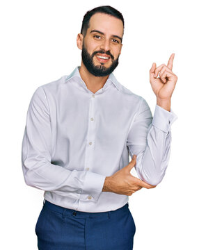 Young man with beard wearing business shirt with a big smile on face, pointing with hand and finger to the side looking at the camera.