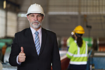 A man in a suit and a hard hat stands in front of a building. He is wearing a tie and a white helmet, show thumb up