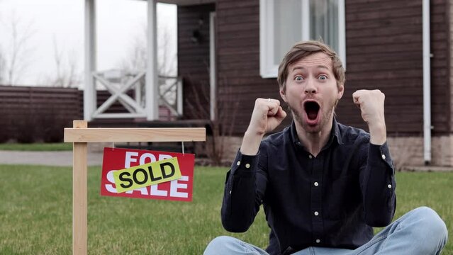 Portrait of excited young man sitting near sold signboard outside house. Happy caucasian man buying new home.