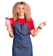 Young blonde woman with curly hair wearing hairdresser apron and holding diploma smiling happy pointing with hand and finger to the side
