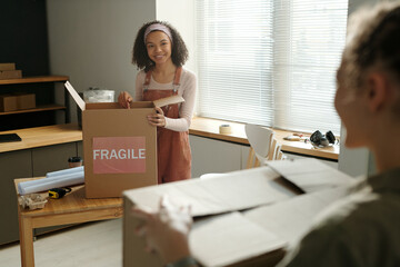 Happy young African American female owner of cafe standing by wooden table and unpacking cardboard...
