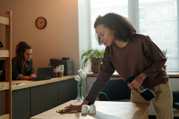 Young African American businesswoman in casualwear wiping table with duster while preparing cafe for opening against girl with laptop - obrazy, fototapety, plakaty