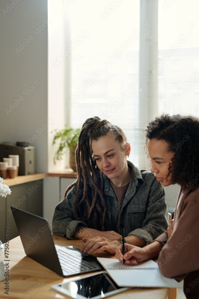 Poster two young intercultural businesswomen looking at laptop screen while one of them making notes in cop