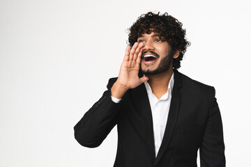 Attractive Hindu young businessman in black tuxedo shouting using hand on mouth gesture isolated over white background. Indian manager telling secret top news