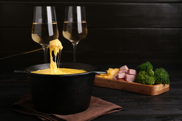 Dipping piece of ham into fondue pot with melted cheese on black wooden table