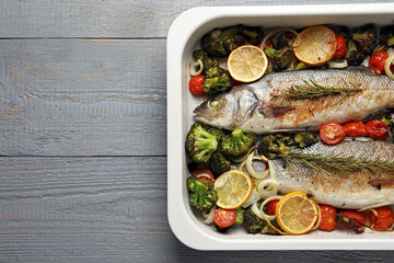 Delicious fish with vegetables and lemon in baking dish on grey wooden table, top view. Space for text