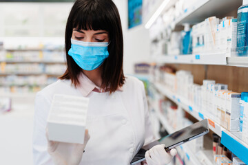 Beautiful pharmacist with protective face mask working and standing in a drug store and doing a...