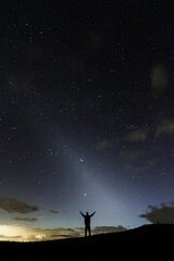 A stargazer having his hands open wide under the night sky  in the middle silhuette showing the zodiacal light in the sky.