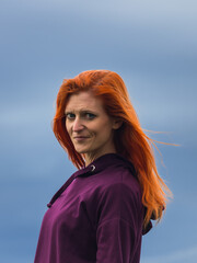 Portrait of redheaded smiling young woman against blue cloud - 775077220