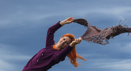Portrait of redheaded young woman playing with blowing scarf in wind against blue cloud - 775077251